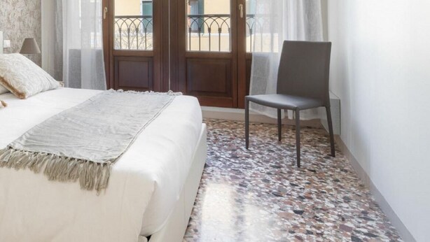 Quarto com cama, uma cadeira e vista para edifícios através de uma porta.