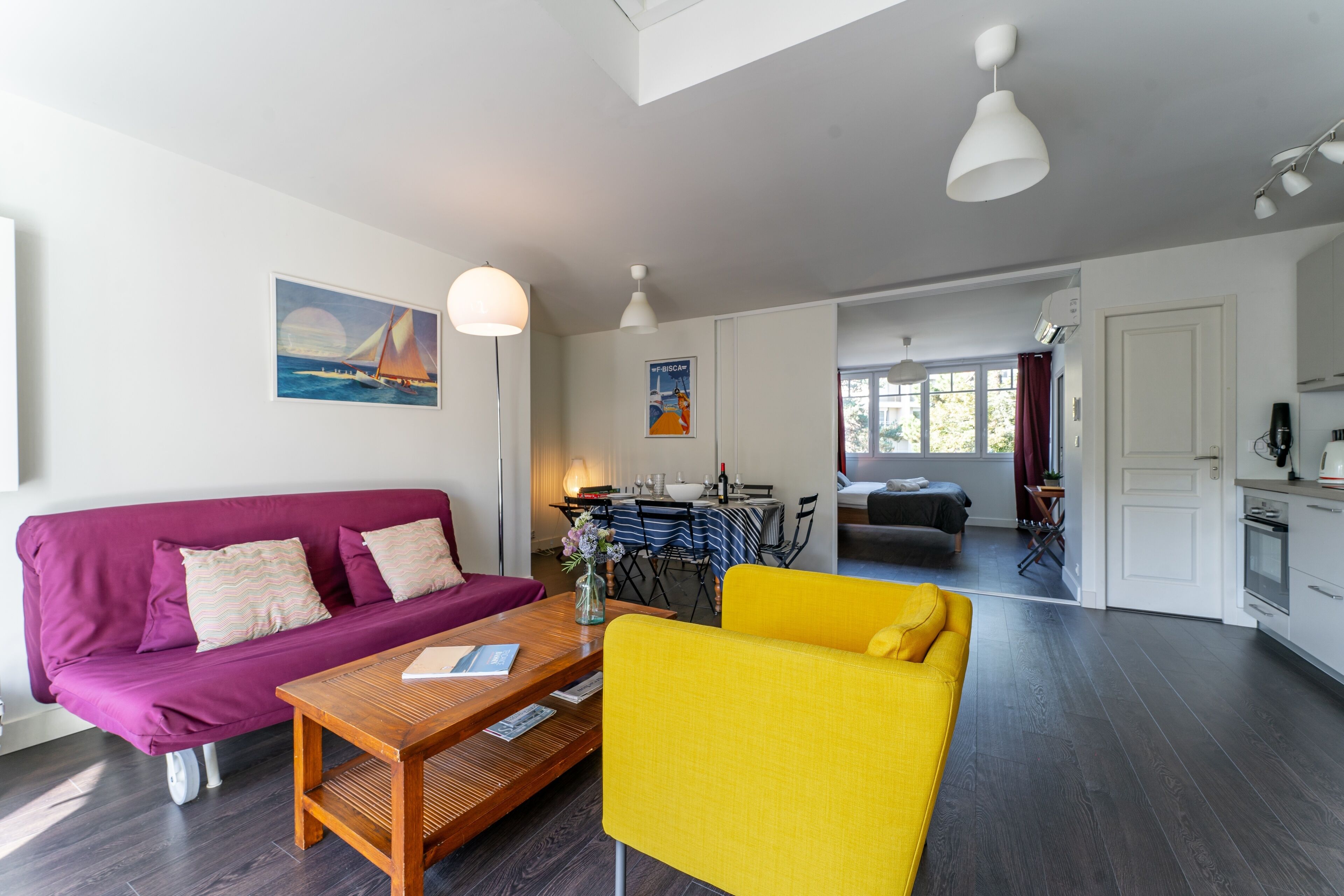 Una sala moderna con un sofá morado, un sillón amarillo y una mesa de centro de madera. Hay una zona de comedor con mantel a rayas y sillas, y una cocina con armarios blancos y una estufa.
