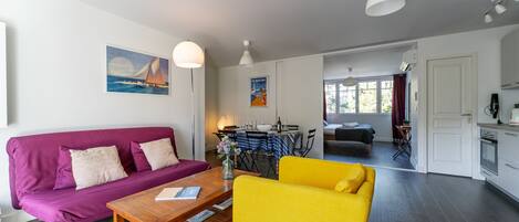 Una sala moderna con un sofá morado, un sillón amarillo y una mesa de centro de madera. Hay una zona de comedor con mantel a rayas y sillas, y una cocina con armarios blancos y una estufa.