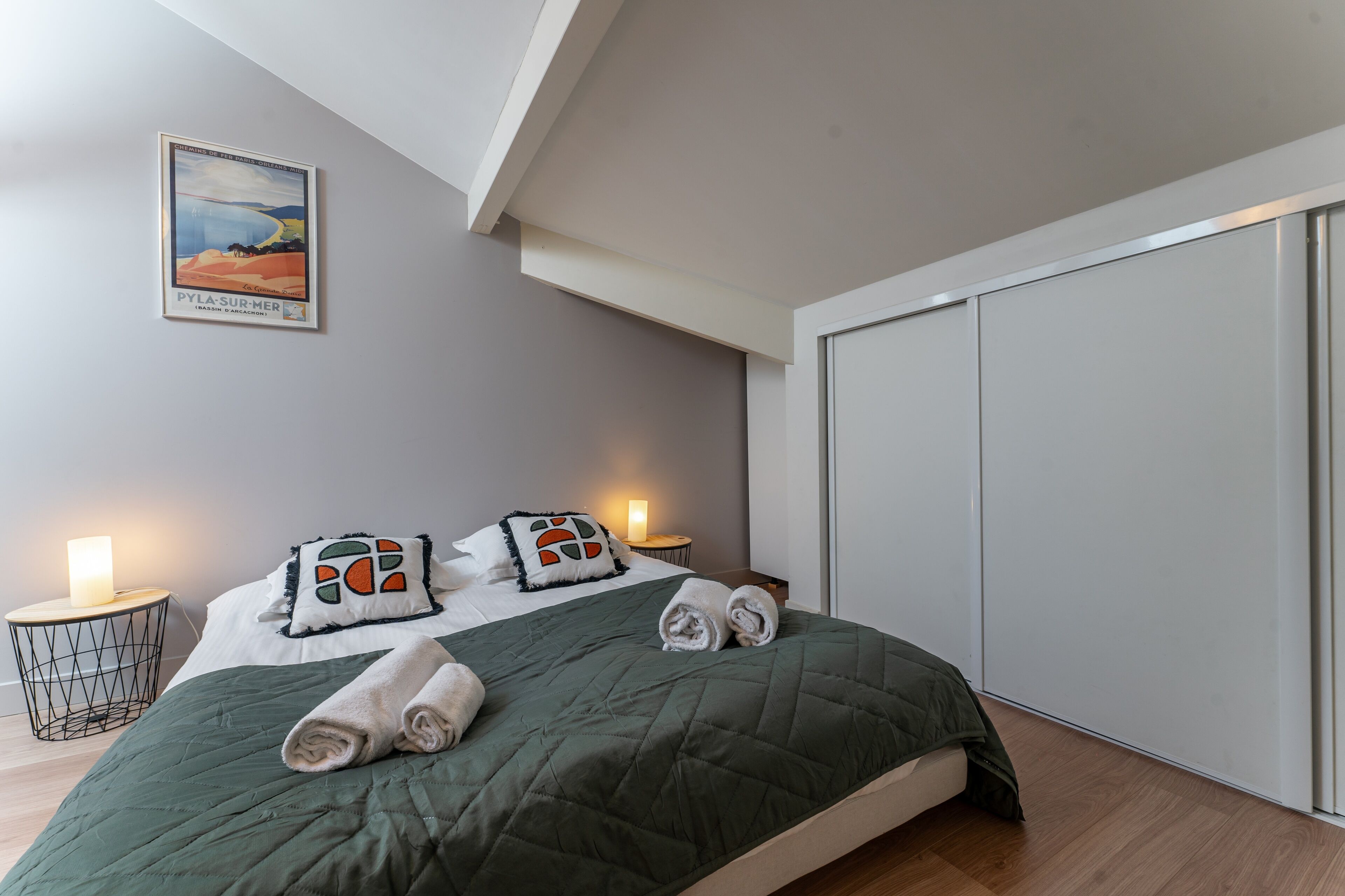 Een moderne slaapkamer met een groen gewatteerde spreidekker, twee kussens met geometrische patronen, een ingelijste foto aan de muur en een inloopkast met schuifdeuren.