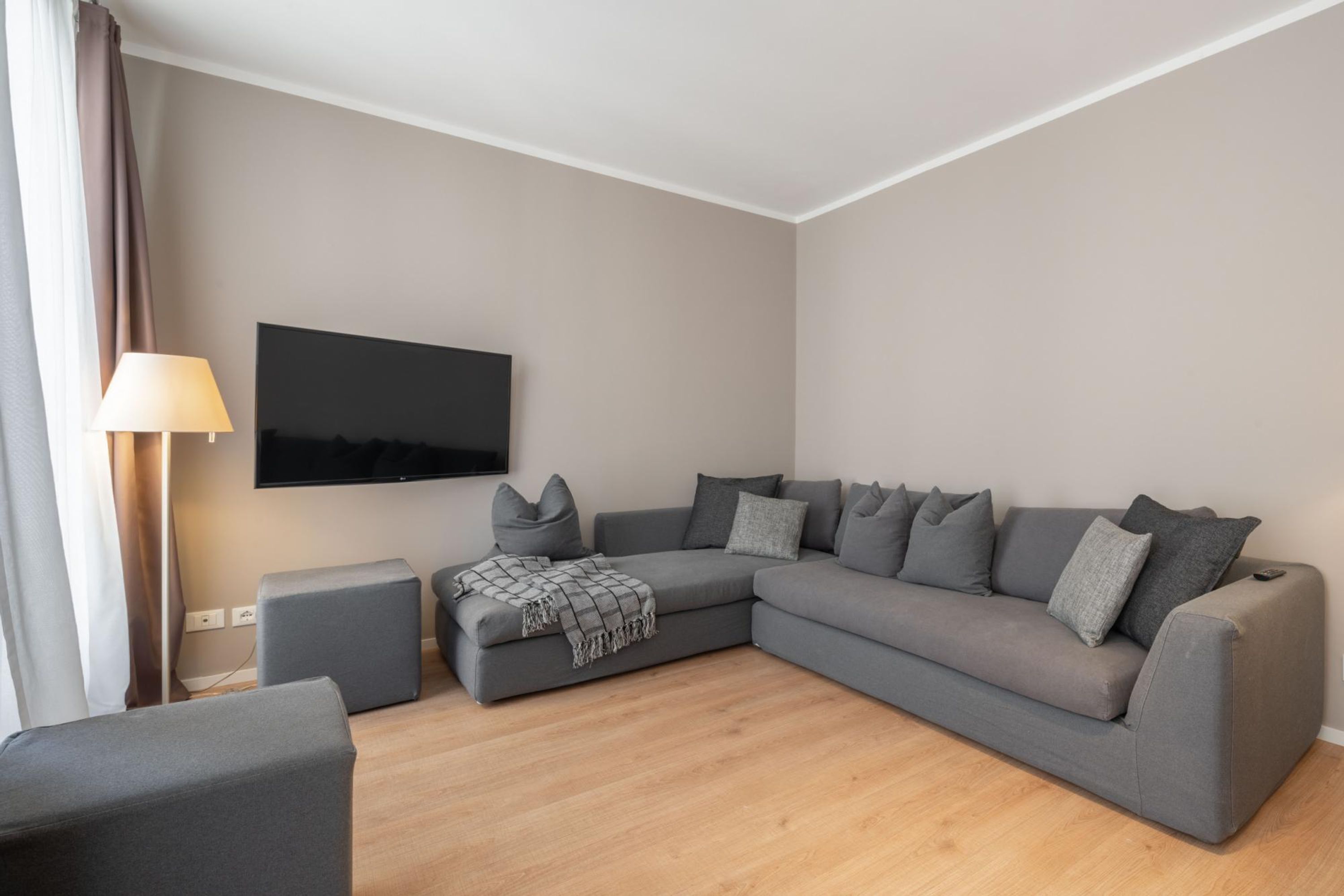 A modern living room with a grey sectional sofa, a small ottoman, and a flat-screen TV mounted on the wall.