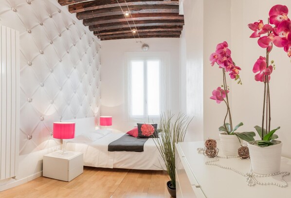 A bedroom with a tufted headboard, a bed with white bedding and pink pillows, a bedside table with a pink lamp, and a window with natural light.
