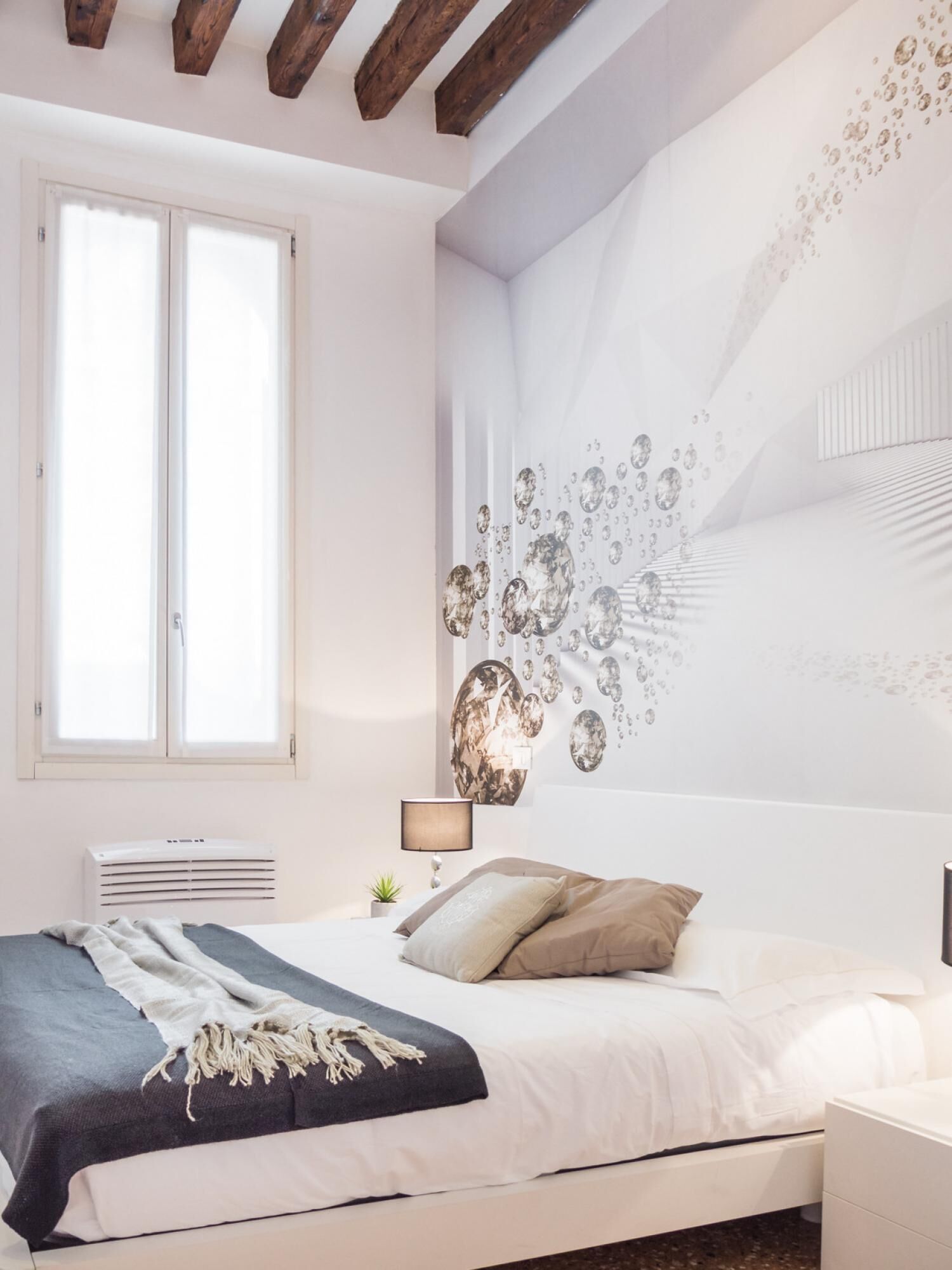 Une chambre à coucher comprenant un lit, un climatiseur fixé au mur, une lampe de chevet et une fenêtre avec des stores.