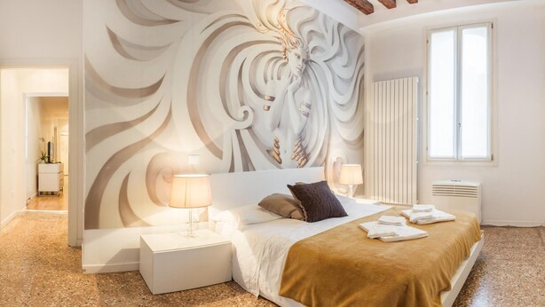 A modern bedroom with a large wall mural, a bed with a brown blanket, two bedside lamps, and a window with blinds. - Rialto Project Apartment 4 by Wonderful Italy (Venice)