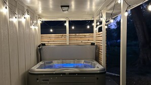 A covered patio area with a hot tub, a table, and chairs. - Cozy Modern Spa ~ Family Fun meets Serene Luxury (Sioux Falls)