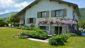 Una casa de dos pisos con techo inclinado, contraventanas de madera y un jardín con diversas plantas y flores.