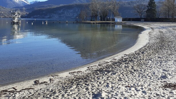 Un lago transparente con orilla de guijarros, un muelle que se adentra en el agua y una cadena montañosa al fondo.