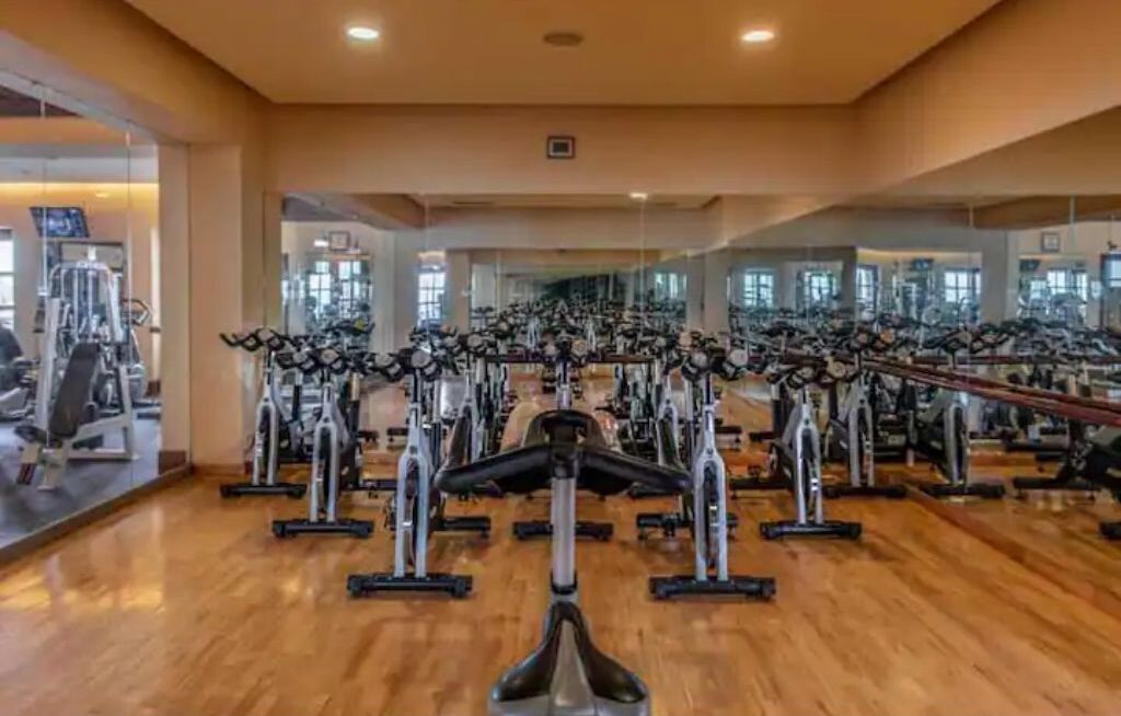 A gym with multiple exercise bikes and cardio machines, wooden flooring, and large windows.