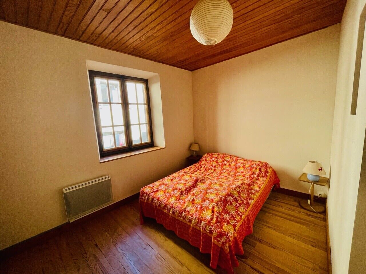 A bedroom with a wooden floor, a bed with a floral bedspread, a window, a radiator, and a bedside lamp.