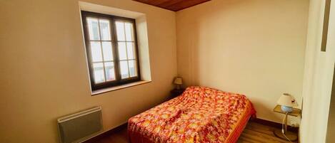 A bedroom with a wooden floor, a bed with a floral bedspread, a window, a radiator, and a bedside lamp.