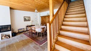 A wooden staircase leading to an upper floor, a dining area with a table and chairs, a fireplace, and a ceiling with wooden beams.