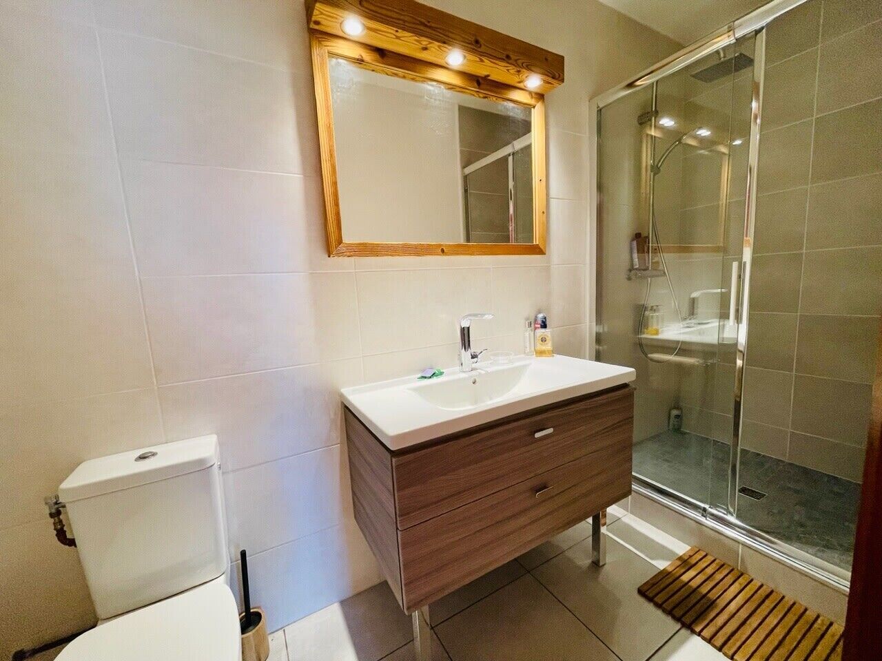 Une salle de bain avec des toilettes blanches, un meuble-lavabo en bois avec un évier blanc et une cabine de douche en verre.