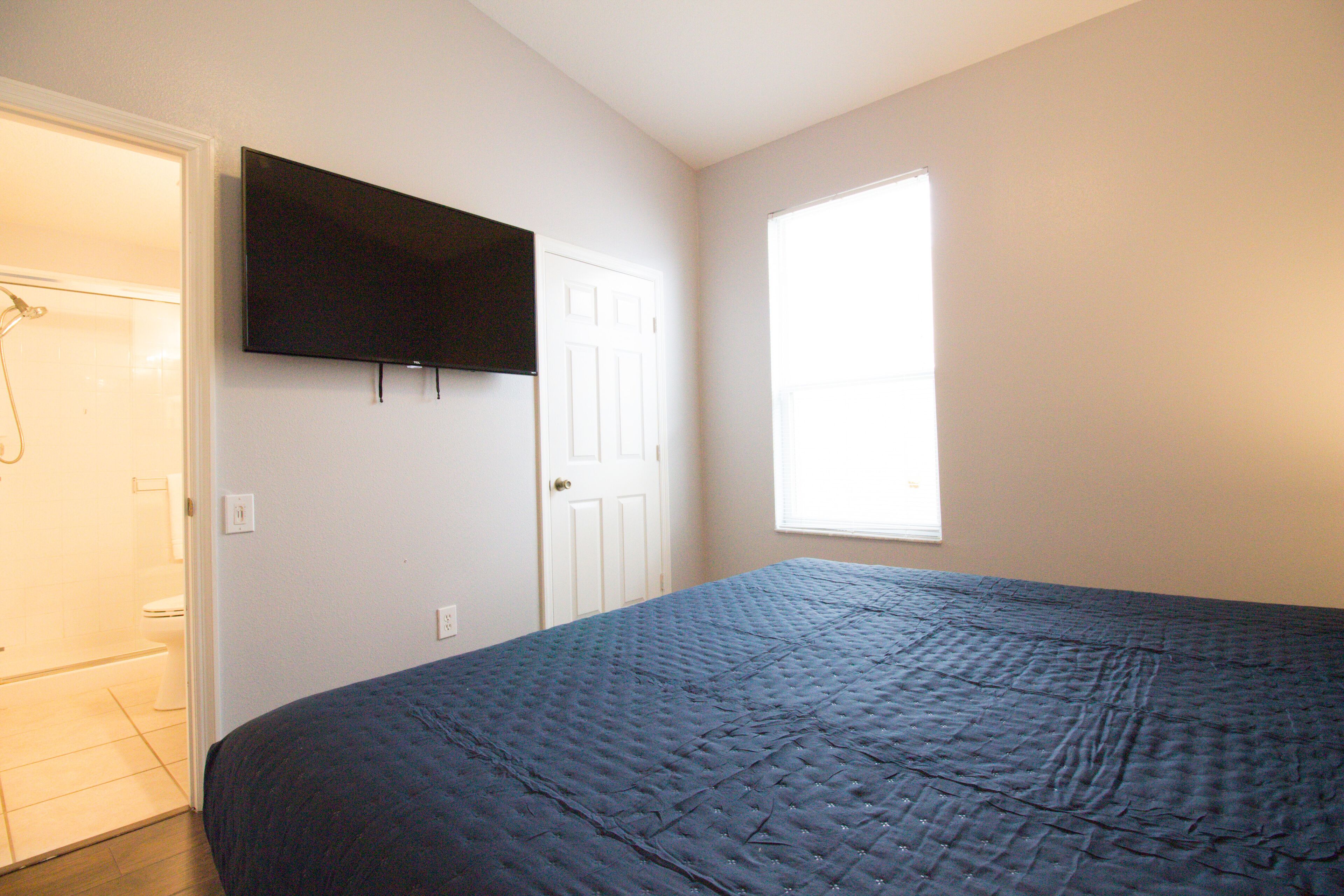 A bedroom with a bed, a television mounted on the wall, a door, and a window.