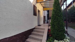 Une entrée d’immeuble avec un escalier couvert, un petit jardin fleuri et un grand arbre à feuilles persistantes.
