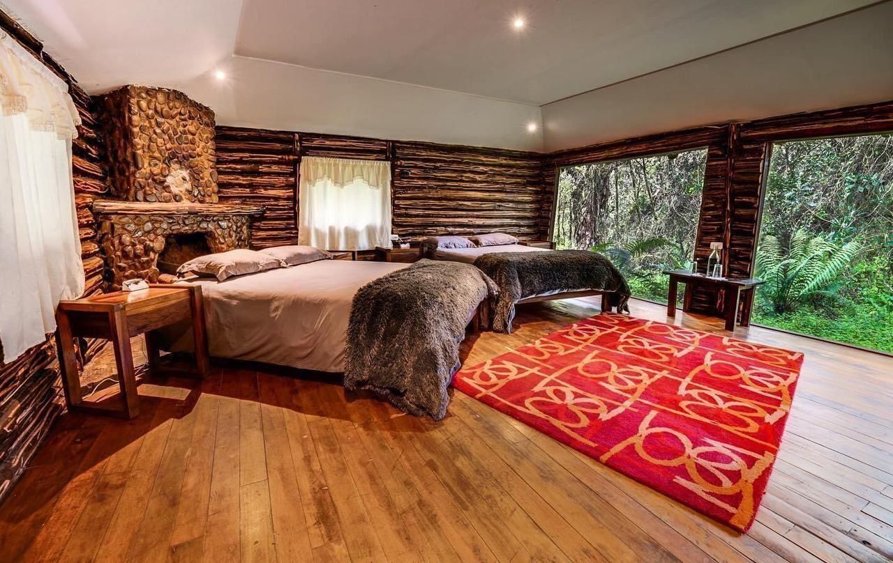 A bedroom with a stone fireplace, two beds, a nightstand, and a large window overlooking a forest.
