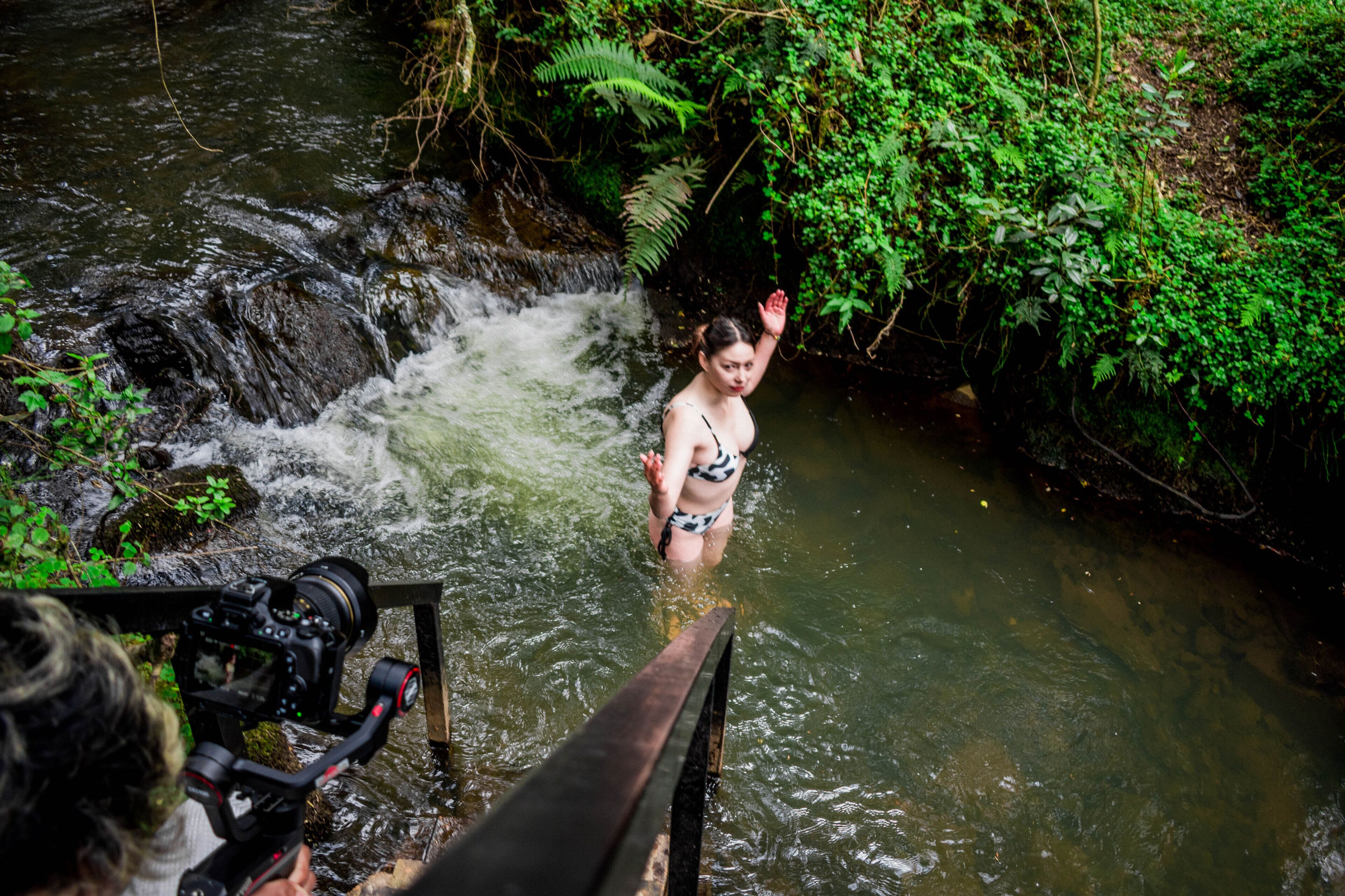 A person in a bikini standing in a stream with a camera on a tripod.