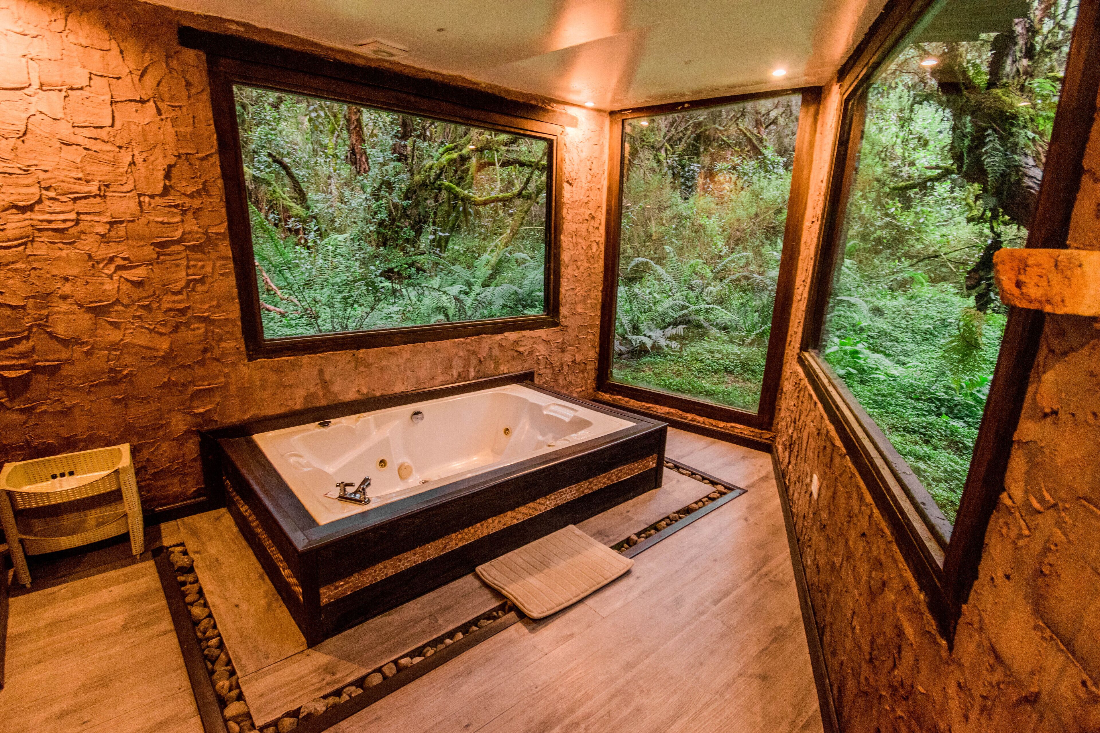 Un baño moderno con una bañera grande, suelo de madera, paredes de piedra y ventanas amplias que dan a un bosque.