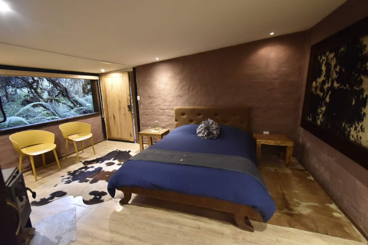 Un dormitorio con una cama grande, cabecero de madera, alfombra de cuero de vaca y una ventana amplia con vista al bosque.