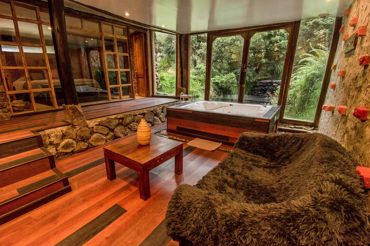 Un cuarto de spa con piso de madera, un ventanal grande, pared de piedra, un banco de madera, una mesa de madera y una bañera amplia.