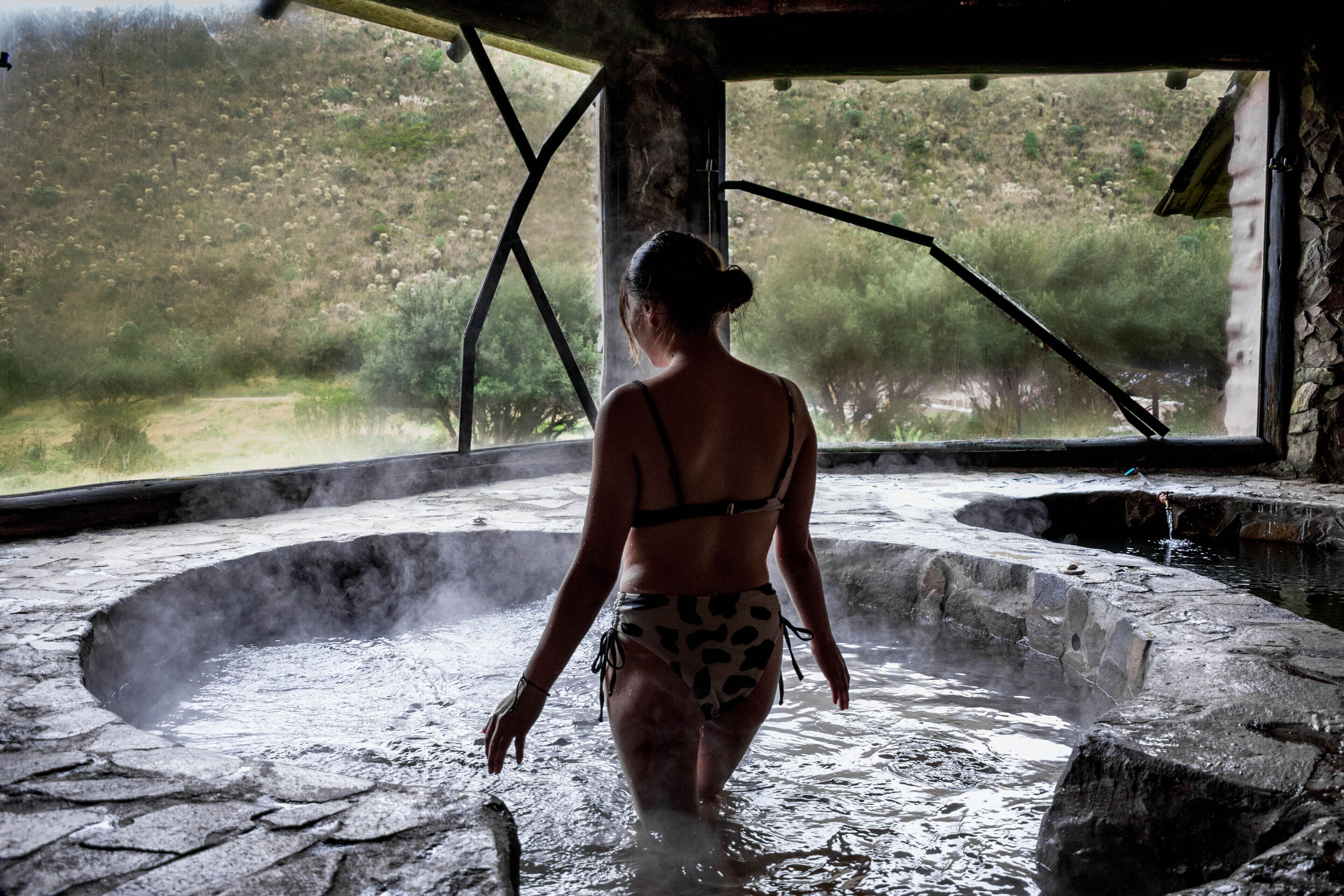 A woman in a bikini walking through a stone hot spring area with steam rising from the water.