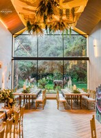 A dining area with a long table set for a meal, large windows offering a view of the outdoors, and a ceiling with hanging plants.