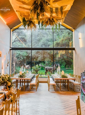 Un comedor con una mesa larga preparada para una comida, ventanas amplias que ofrecen vistas al exterior y un techo decorado con plantas colgantes.
