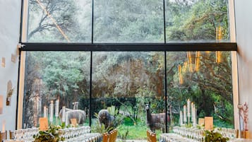 Un comedor con un gran ventanal que da a un jardín con llamas, sillas de madera y mesas iluminadas con velas.