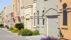 Une rangée de maisons aux façades uniformes, de couleur beige, avec des toits recouverts de tuiles rouges, alignées le long d’une rue bordée d’un trottoir et de parterres de fleurs.
