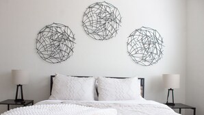 Une chambre à coucher avec un lit, du linge de lit blanc, deux tables de chevet avec des lampes et trois œuvres d’art abstraites accrochées au mur.