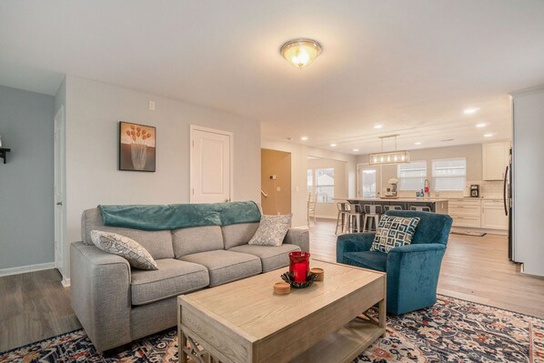 A modern living room with a grey sofa, a blue armchair, and a wooden coffee table. A kitchen is visible in the background with white cabinets and a dining area. - Eco Friendly Getaway (Lowell)