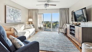Un salon moderne comprenant un canapé, un fauteuil et un meuble de rangement pour appareils électroniques, avec une grande télévision et un ventilateur de plafond.