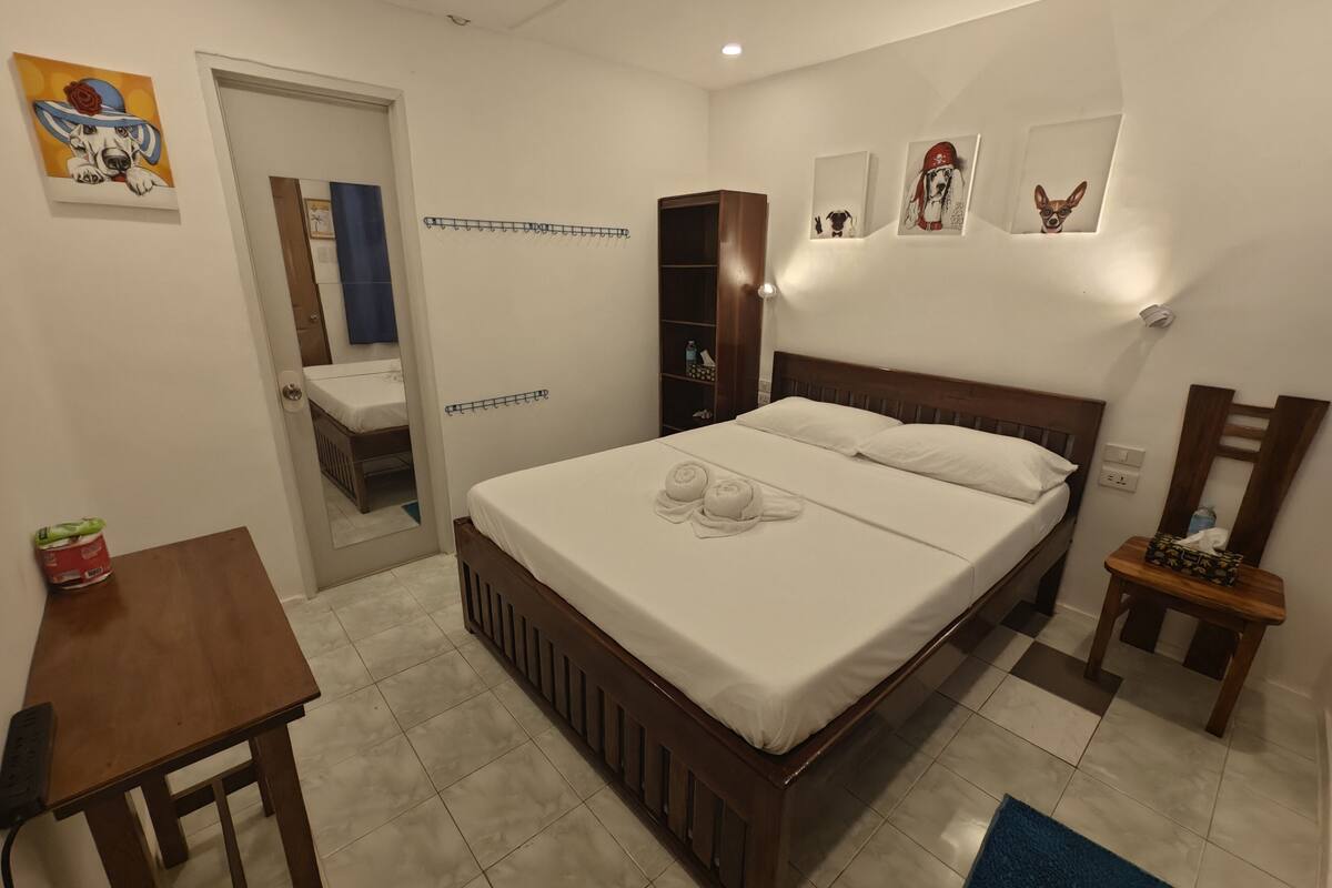 A bedroom with a wooden bed, white linens, a small table, a chair, and artwork on the walls.