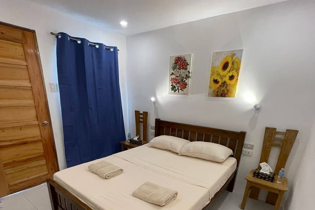 A bedroom with a wooden bed, white bedding, a blue curtain, a small bedside table with a lamp, and two wall-mounted paintings.