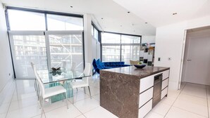 A modern kitchen with a marble island, large windows, and a dining area with a glass table and chairs. - River Side View Luxury Penthouse Apartment (London)