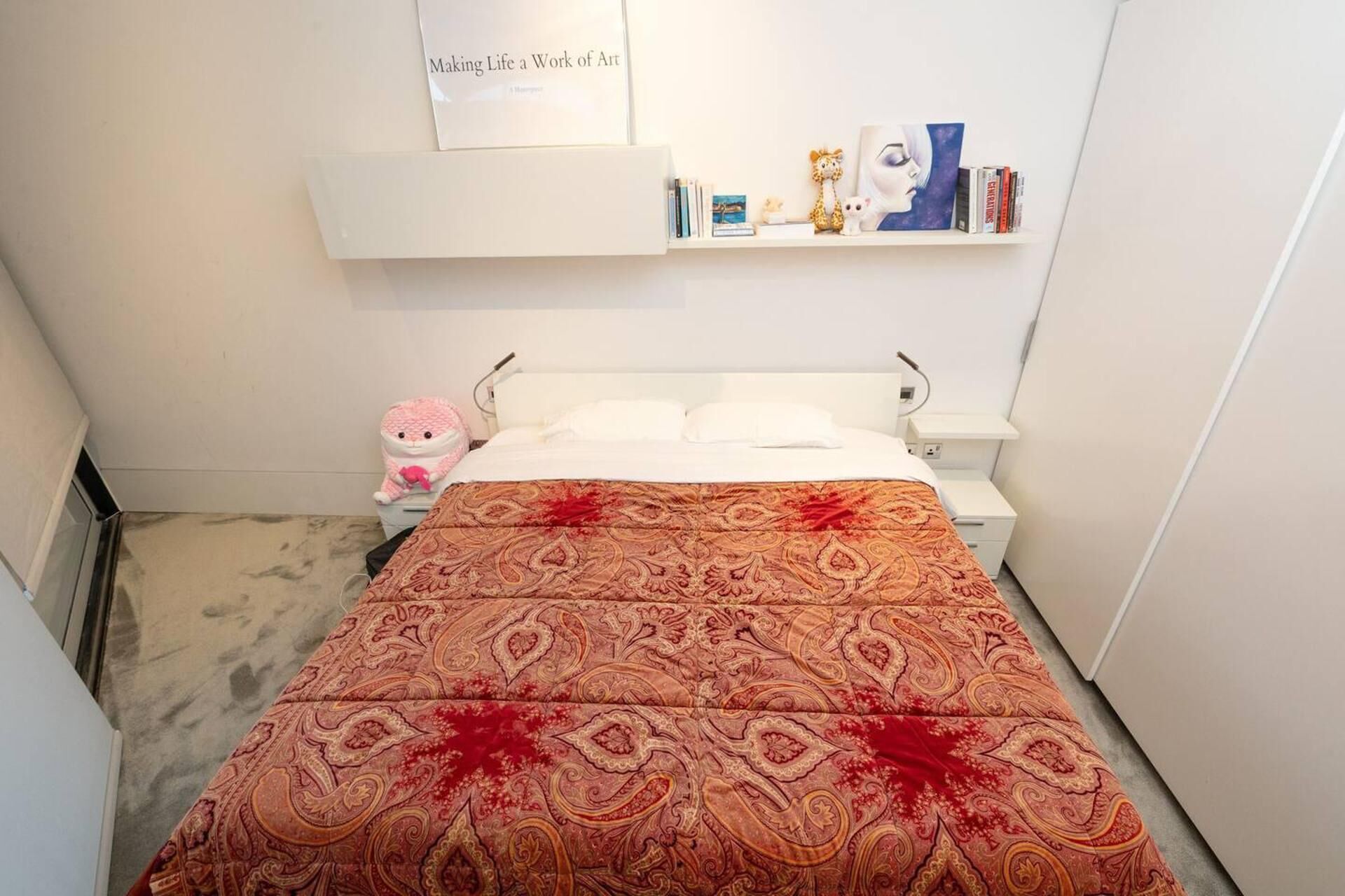 A neatly made bed with a patterned bedspread, a bedside table with a lamp, a shelf with books and decorative items, and a small stuffed toy on the floor.