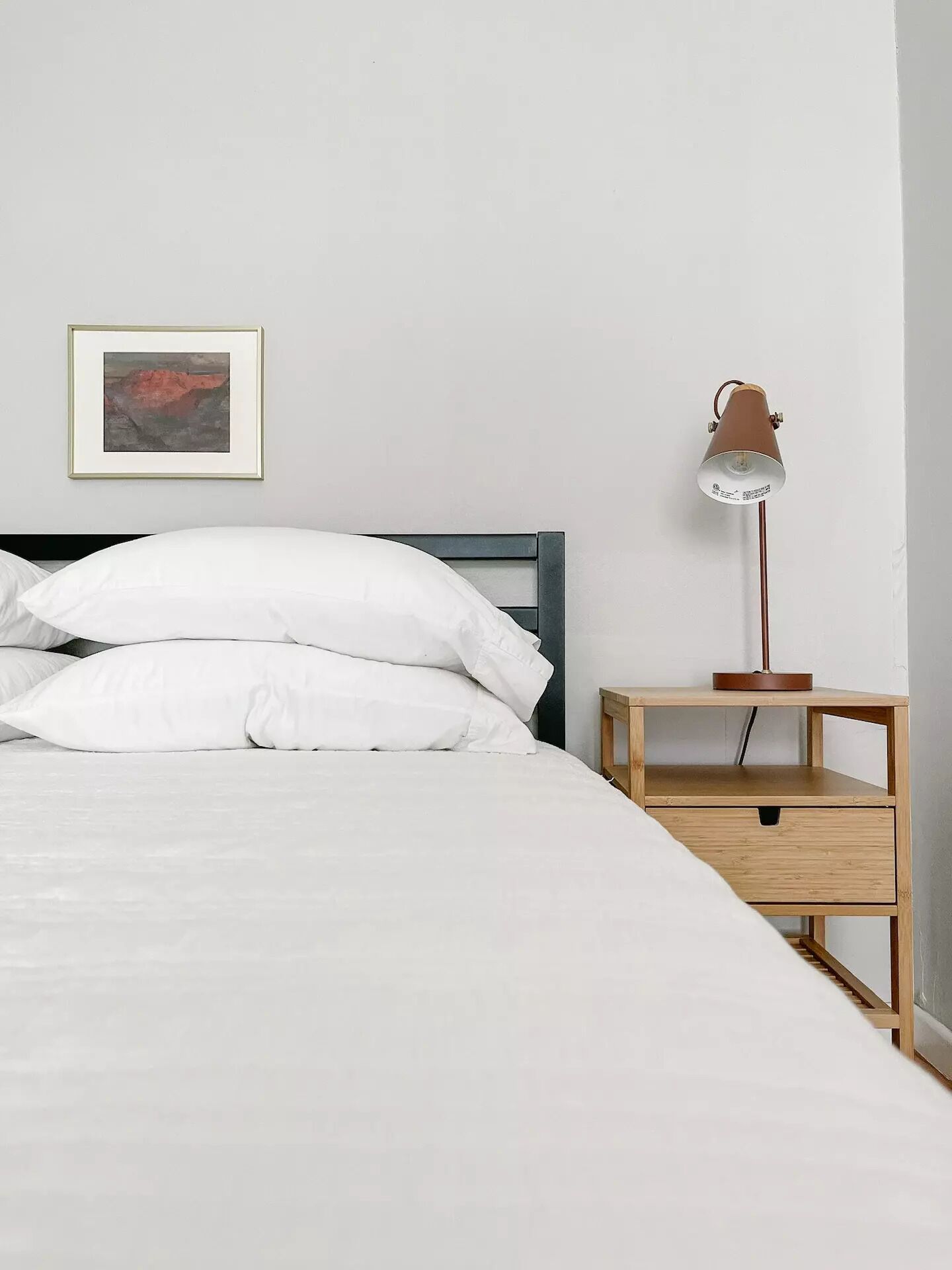 Una cama bien hecha con ropa de cama y almohadas blancas, un cuadro abstracto enmarcado en la pared, y una mesita de noche de madera con una lámpara.