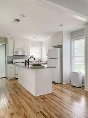 A modern kitchen with white cabinetry, a central island, a microwave, a refrigerator, and a window with blinds. - Boutique home near ACU (Abilene)