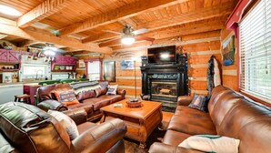 Un acogedor interior de cabaña con sofás de cuero, chimenea, paredes de madera y una zona de cocina.
