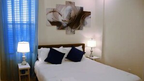 A neatly made bed with white linens and blue pillows, a wall-mounted abstract painting, and two bedside lamps on either side. - Spacious 5BR/5BA Group Stay w/Game Room + Parking, Sleeps 12, Central Location (Pittsburgh)