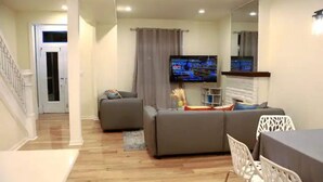 A living room with a grey sofa, a TV, and a dining area with a table and chairs. - Spacious 5BR/5BA Group Stay w/Game Room + Parking, Sleeps 12, Central Location (Pittsburgh)