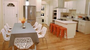 A modern kitchen with white cabinetry, a stainless steel refrigerator, a gas stove, and a dining area with white chairs and a table. - Spacious 5BR/5BA Group Stay w/Game Room + Parking, Sleeps 12, Central Location (Pittsburgh)