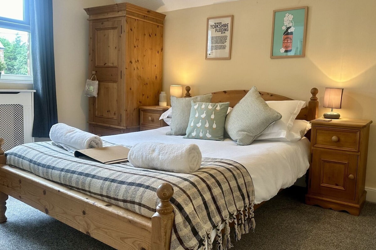 A cozy bedroom with a wooden bed, a nightstand, a wooden wardrobe, and a framed recipe on the wall.
