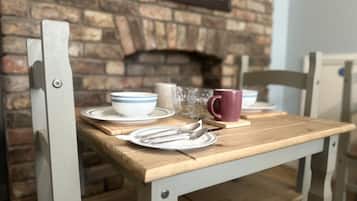 A wooden dining table with plates, cutlery, and a mug set against a brick wall with a fireplace.