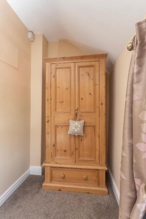 Guarda-roupa de madeira com um pequeno objeto decorativo no topo, situado num canto de um quarto com paredes bege e chão coberto de carpete.
