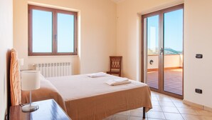 A bedroom with a bed, a nightstand, a lamp, a chair, a radiator, and a sliding glass door leading to a balcony with a view.