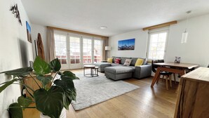 Un salon spacieux avec un canapé gris, une table basse ronde et une table à manger avec des chaises. Un grand pot de plantes se trouve dans un coin, et on peut admirer la vue sur l’extérieur à travers les portes-fenêtres coulissantes.