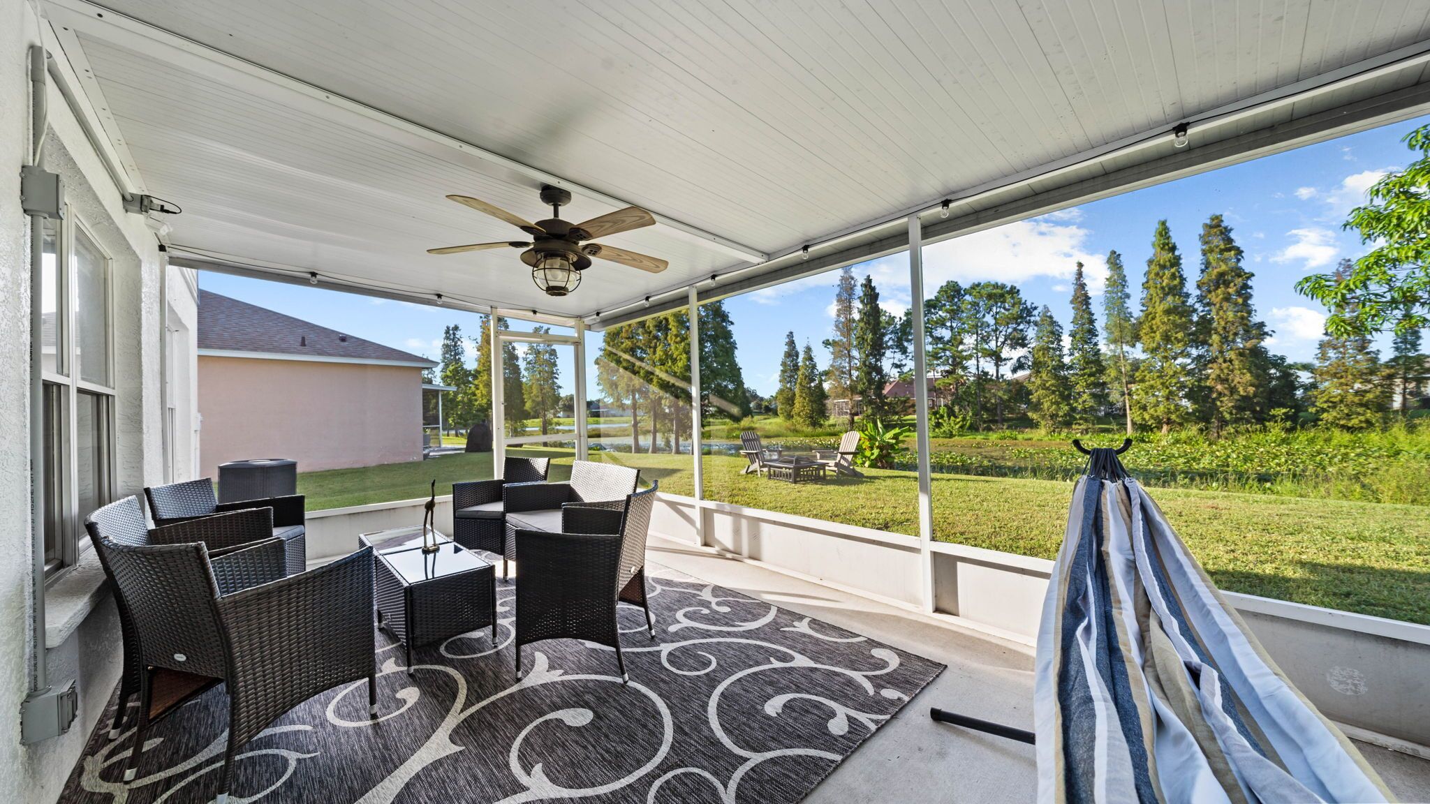 Eine überdachte Terrasse mit Korbmöbeln, einem Deckenventilator und Blick auf eine begrünte Fläche mit Bäumen.