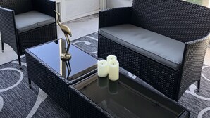 En udendørs siddeplads overdækket med fletningsmøbler og et glastbord, med udsigt over en sø og træer.