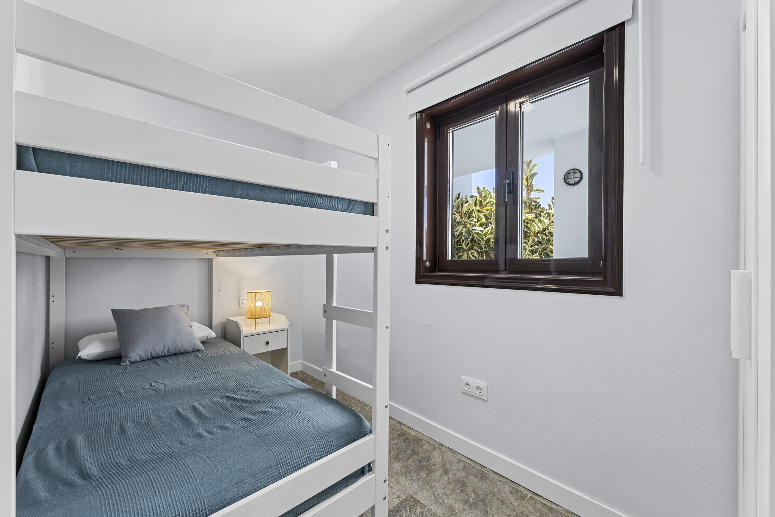 A bunk bed room with a window, a bedside table with a lamp, and a view of greenery outside.
