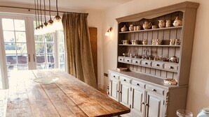 A dining area with a wooden table, a cabinet with kitchenware, and a window with curtains. - French Vintage Cottage in the Heart of the Fens (Spalding)